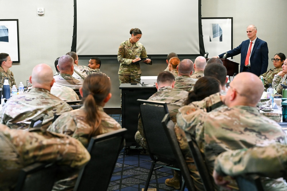 A military briefing or training session is taking place in a conference room setting. A female service member in OCP (Operational Camouflage Pattern) uniform stands at the front reading from a document, while a civilian male in a suit stands at a podium to the right. Approximately 20 uniformed military personnel are seated at tables facing the presenters.