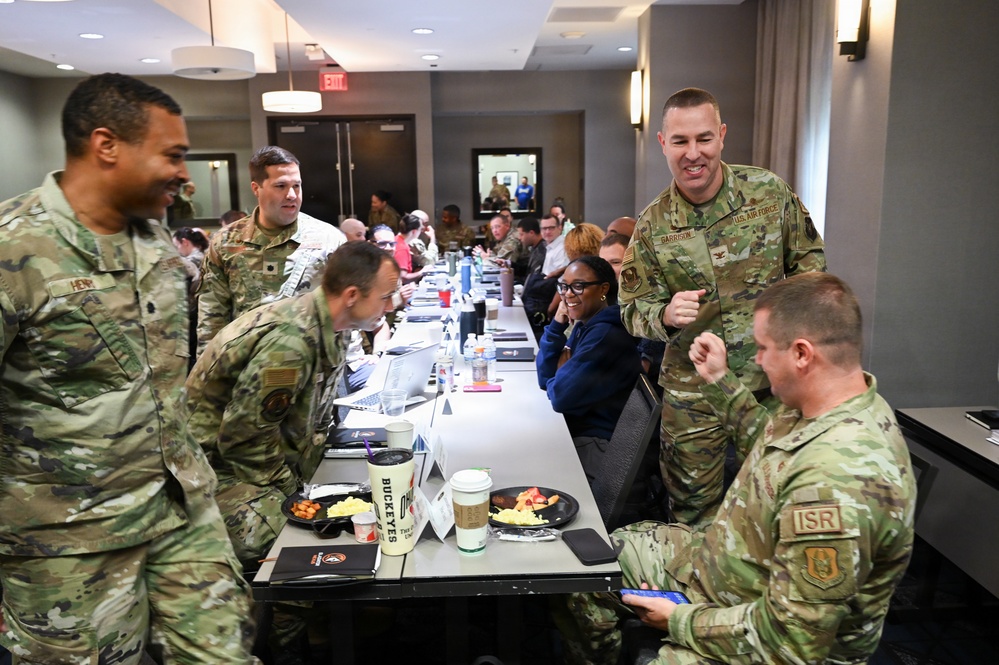 A group of U.S. Air Force personnel in OCP (Operational Camouflage Pattern) uniforms are gathered in what appears to be a hotel or conference room setting during a meal or briefing event. Several uniformed members are standing and conversing while others are seated at a long table with food plates and beverages. The atmosphere appears informal and social, suggesting a working breakfast or leadership gathering.