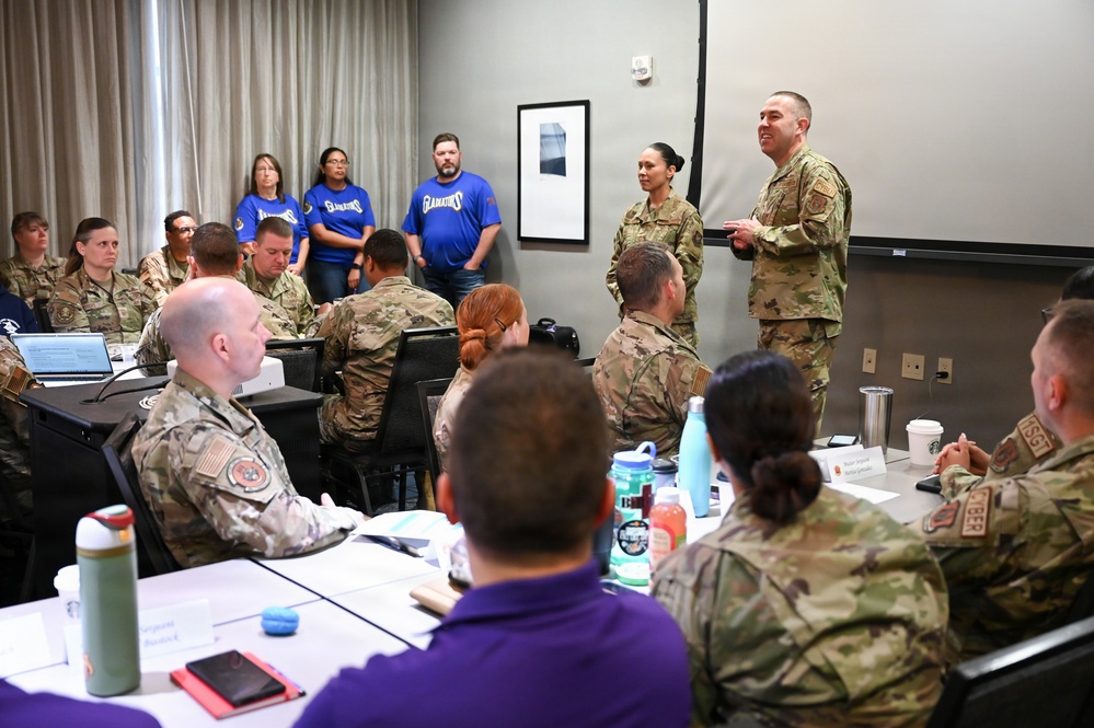 A military briefing or training session is taking place in a conference room, with a male service member in OCP (Operational Camouflage Pattern) uniform appearing to address the group. The majority of attendees are also in OCP uniforms, seated at tables, while several civilians in blue 'Gladiators' t-shirts stand along the back wall. The setting appears to be a formal military professional development or community engagement event.