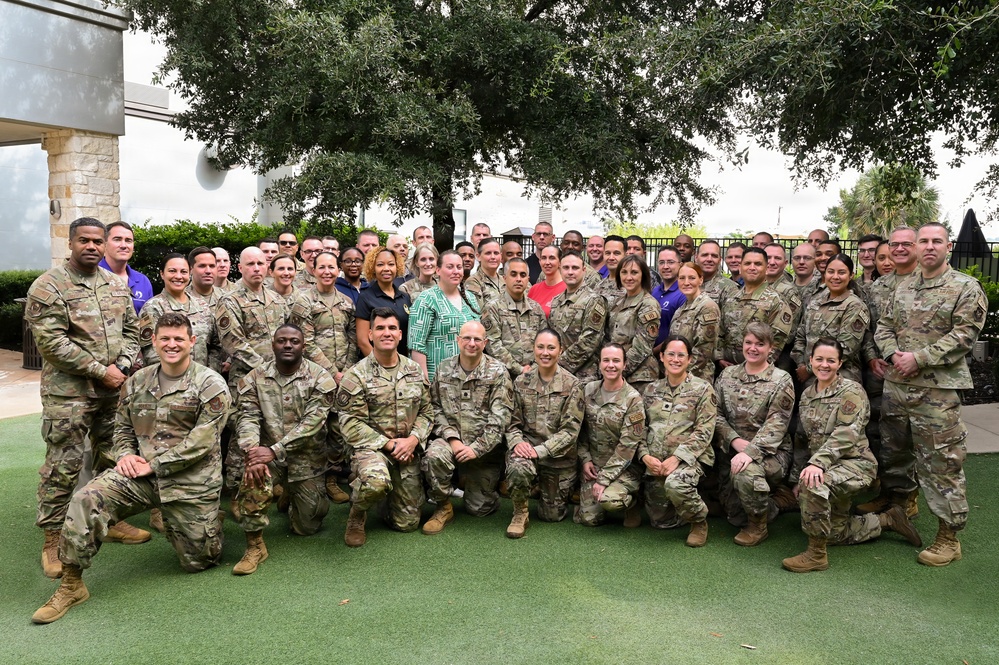 A large group photograph of approximately 50-60 military personnel, predominantly in U.S. Air Force/Space Force OCP (Operational Camouflage Pattern) uniforms, posed outdoors. A small number of civilians in casual attire are interspersed among the group. The photo appears to be a formal unit or conference group photo taken on a manicured outdoor patio area.