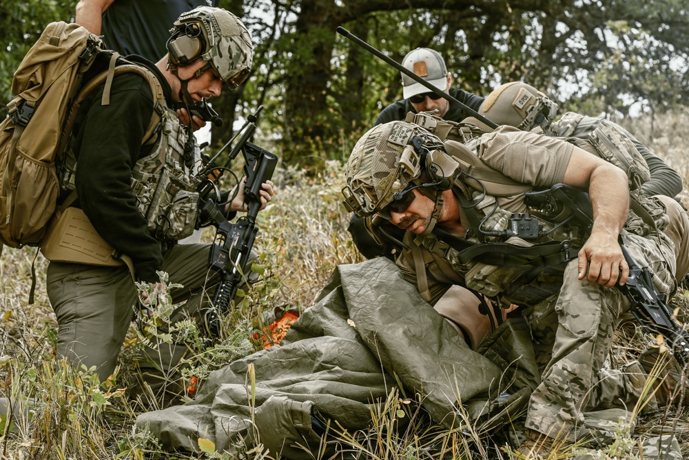 The image depicts multiple individuals in military or tactical gear conducting what appears to be a casualty evacuation or combat medical training exercise in an outdoor, vegetated environment. One person is being lifted or assisted from the ground while wrapped in an olive-drab tarp or emergency blanket. The scene involves at least three armed personnel in full tactical kit alongside one individual in civilian attire.