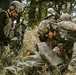 The image depicts multiple individuals in military or tactical gear conducting what appears to be a casualty evacuation or combat medical training exercise in an outdoor, vegetated environment. One person is being lifted or assisted from the ground while wrapped in an olive-drab tarp or emergency blanket. The scene involves at least three armed personnel in full tactical kit alongside one individual in civilian attire.