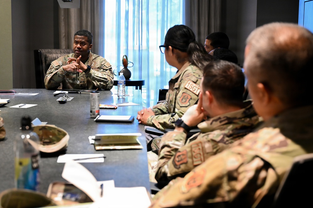 A military briefing or meeting is taking place around a conference table, with one individual in OCP (Operational Camouflage Pattern) uniform appearing to lead the discussion from the head of the table. Several other uniformed personnel are seated along the side of the table, listening attentively. The setting appears to be an indoor conference or meeting room with natural light from curtained windows.