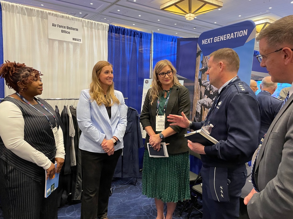 A group of five individuals are gathered at the Air Force Uniform Office booth (MB522) at what appears to be a military or defense conference/expo. The group includes two uniformed Air Force officers and three civilians, engaged in conversation. A 'Next Generation' display booth is visible in the background alongside clothing racks with dark uniforms.