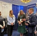 A group of five individuals are gathered at the Air Force Uniform Office booth (MB522) at what appears to be a military or defense conference/expo. The group includes two uniformed Air Force officers and three civilians, engaged in conversation. A 'Next Generation' display booth is visible in the background alongside clothing racks with dark uniforms.