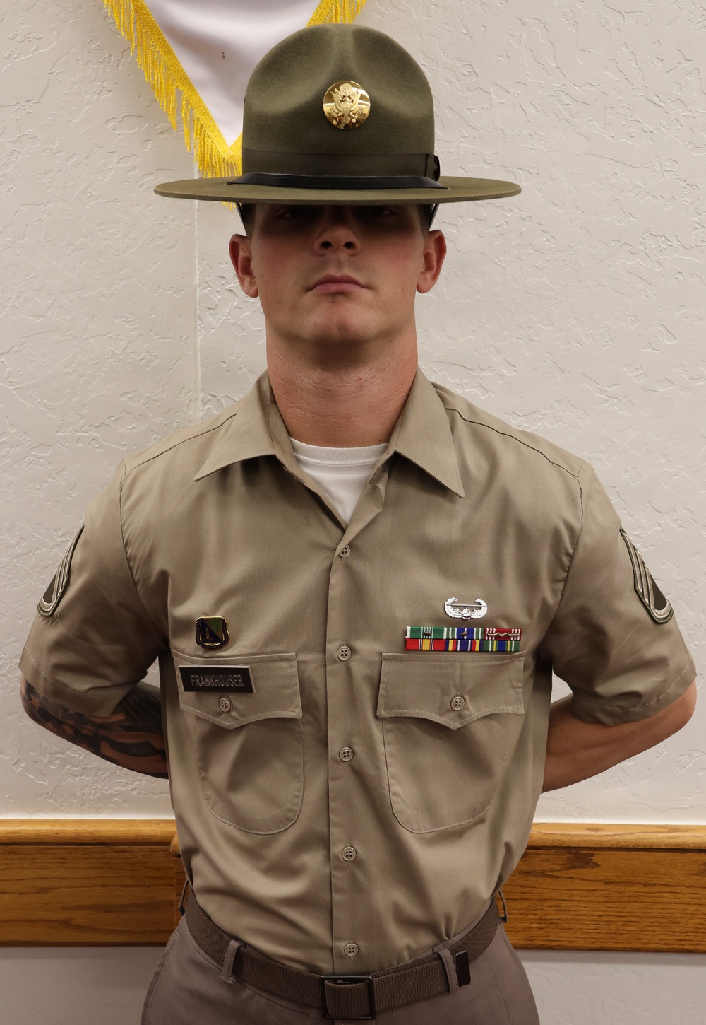 A U.S. Army soldier in Class B uniform (tan short-sleeve shirt) stands at parade rest against a white textured wall. The soldier wears a Drill Sergeant hat (campaign cover) with brass insignia and displays multiple awards and badges on the uniform. A name tape reading 'FRANKHOUSER' is visible on the left chest pocket.