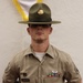 A U.S. Army soldier in Class B uniform (tan short-sleeve shirt) stands at parade rest against a white textured wall. The soldier wears a Drill Sergeant hat (campaign cover) with brass insignia and displays multiple awards and badges on the uniform. A name tape reading 'FRANKHOUSER' is visible on the left chest pocket.