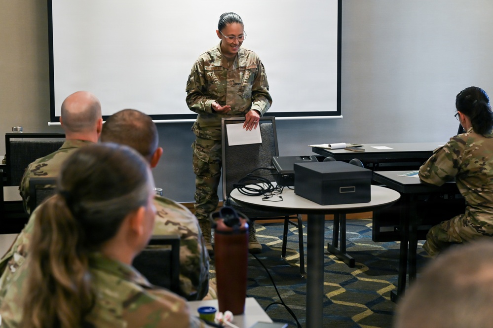A military service member in OCP (Operational Camouflage Pattern) uniform is conducting a briefing or training session in a conference room setting. She is standing at the front of the room holding papers, addressing a small group of fellow service members who are seated at tables. A projection screen is visible behind her, and a projector sits on a nearby table.