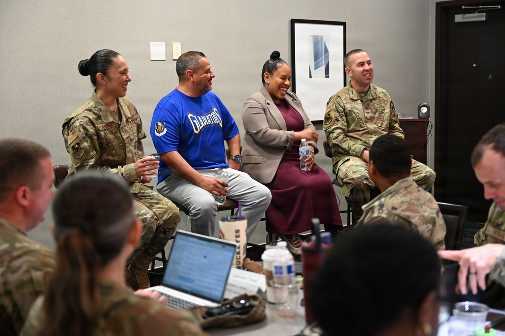 A group of U.S. military personnel in OCP (Operational Camouflage Pattern) uniforms are gathered in a conference or meeting room setting. Four individuals sit at the front as a panel — three in military uniform and one civilian man wearing a blue 'Gladiators' t-shirt, alongside a civilian woman in a blazer and burgundy dress. The atmosphere appears relaxed and positive, with visible laughter and engagement.