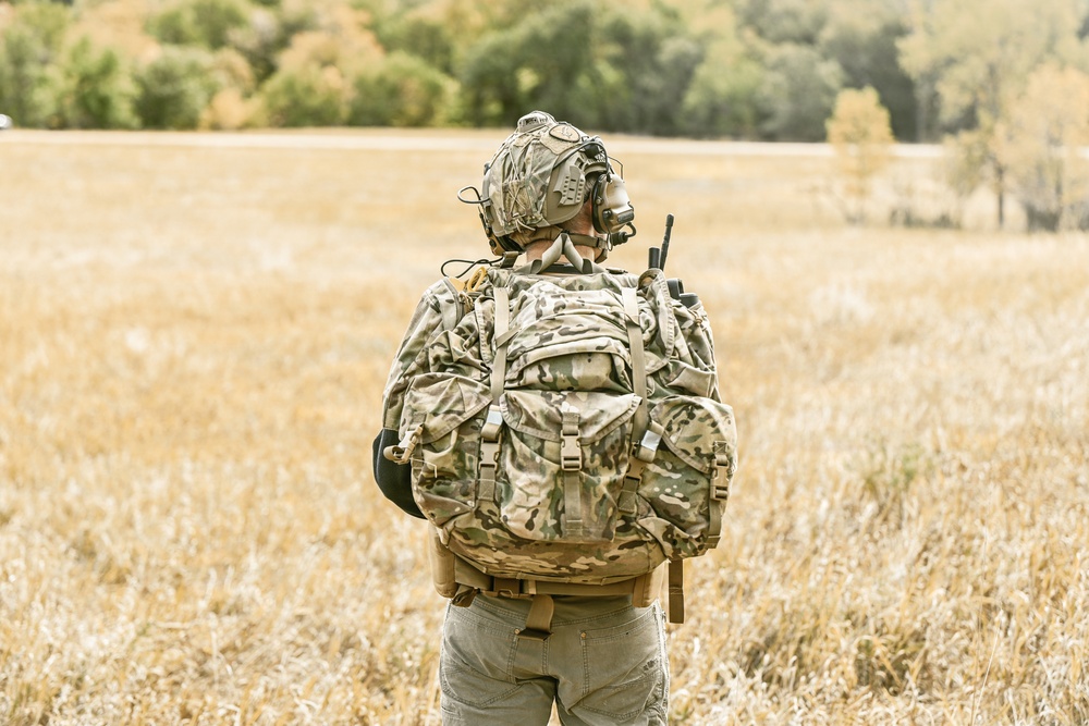 A soldier or tactical operator is seen from behind, standing in an open field of dry golden grass. The individual is fully equipped with military-grade gear including a camouflage uniform, tactical backpack, helmet with communications equipment, and a radio antenna visible over the right shoulder. The background features a treeline with autumn foliage under an overcast sky.