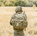 A soldier or tactical operator is seen from behind, standing in an open field of dry golden grass. The individual is fully equipped with military-grade gear including a camouflage uniform, tactical backpack, helmet with communications equipment, and a radio antenna visible over the right shoulder. The background features a treeline with autumn foliage under an overcast sky.