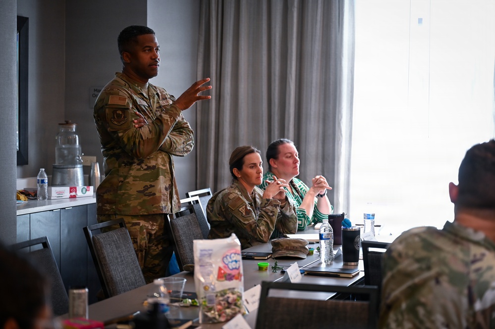 A military briefing or meeting is taking place in what appears to be a conference room or hotel meeting space. A standing male service member in OCP (Operational Camouflage Pattern) uniform is speaking and gesturing while others seated at the table listen attentively. The group includes both uniformed military personnel and at least one civilian participant.