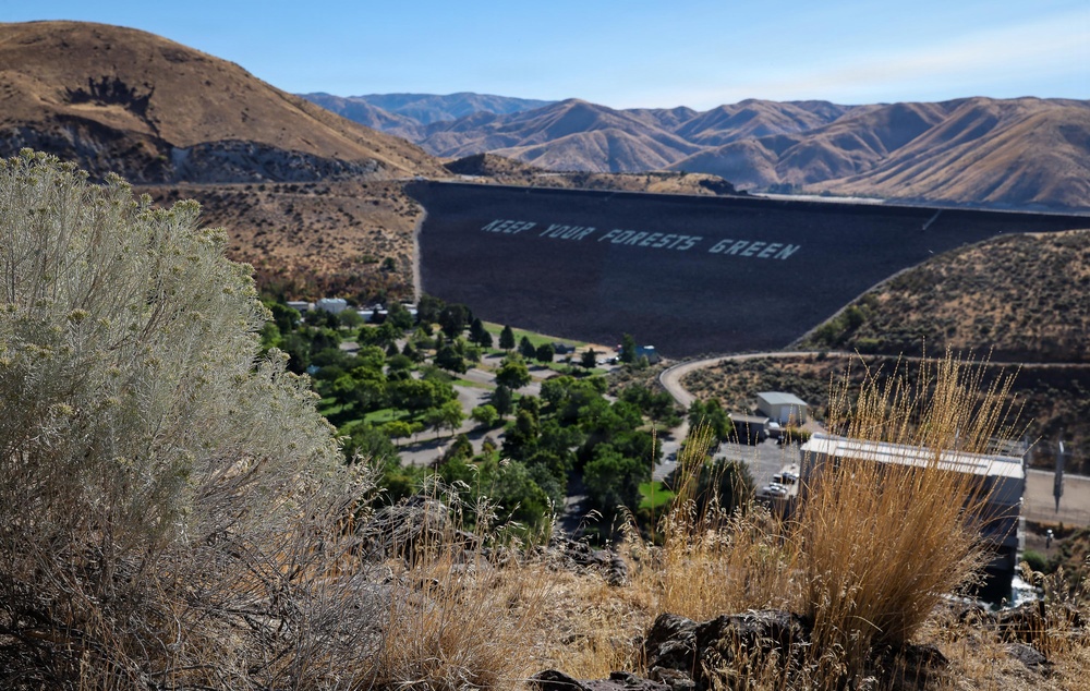 An elevated view of a large earthen dam with the message 'KEEP YOUR FORESTS GREEN' painted in large white letters on its dark face. Below the dam, a green recreational area with trees, parking lots, and facilities contrasts sharply with the surrounding dry, brown hillsides. The arid landscape of rolling mountains stretches into the background under a clear blue sky.