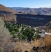 An elevated view of a large earthen dam with the message 'KEEP YOUR FORESTS GREEN' painted in large white letters on its dark face. Below the dam, a green recreational area with trees, parking lots, and facilities contrasts sharply with the surrounding dry, brown hillsides. The arid landscape of rolling mountains stretches into the background under a clear blue sky.