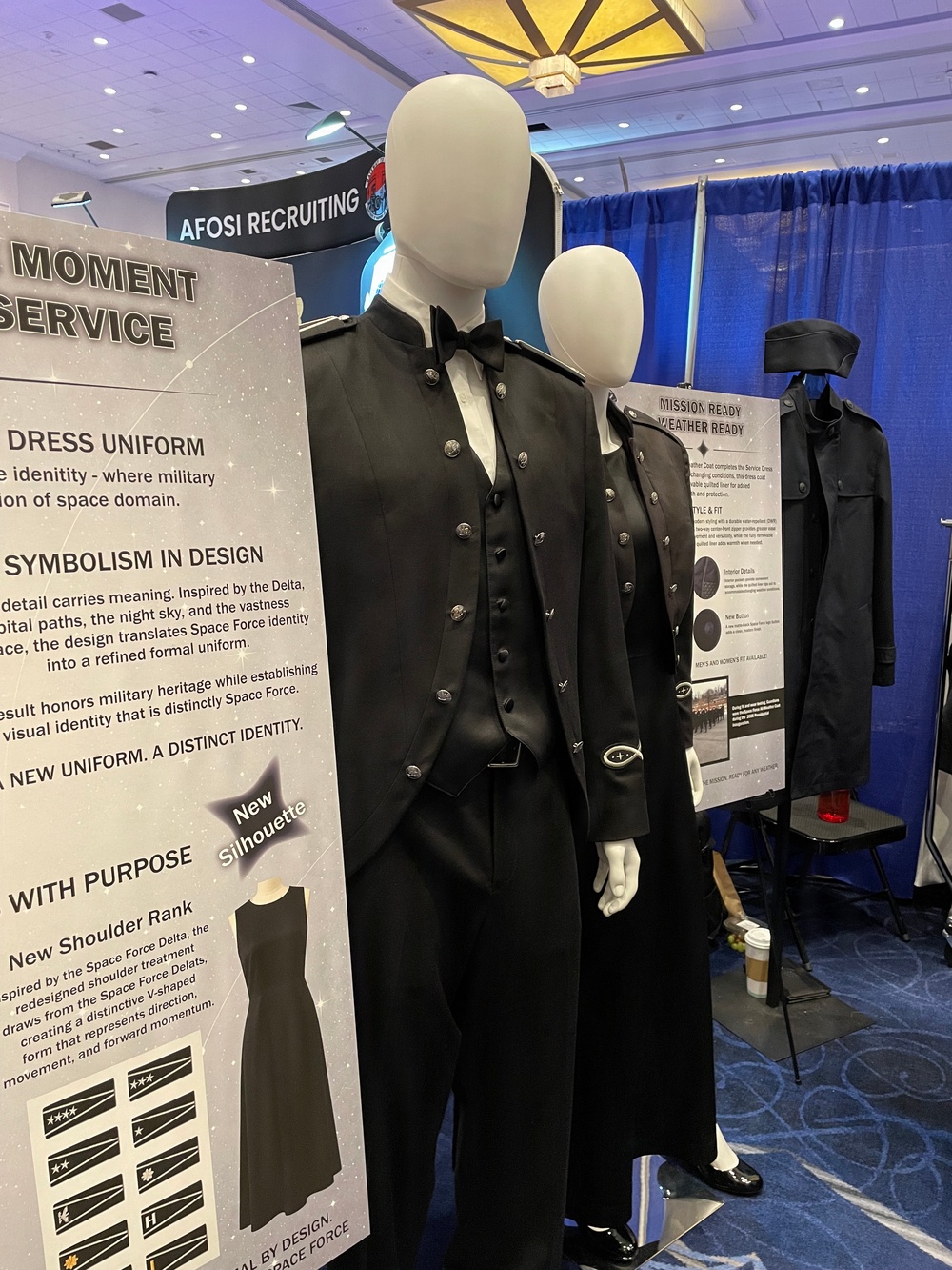 This image shows a military uniform display booth, specifically featuring U.S. Space Force dress uniforms on mannequins at what appears to be a recruiting or trade show event. The display includes informational signage describing the design symbolism, new silhouette, and shoulder rank insignia of the Space Force dress uniform. An 'AFOSI RECRUITING' banner is visible in the background.