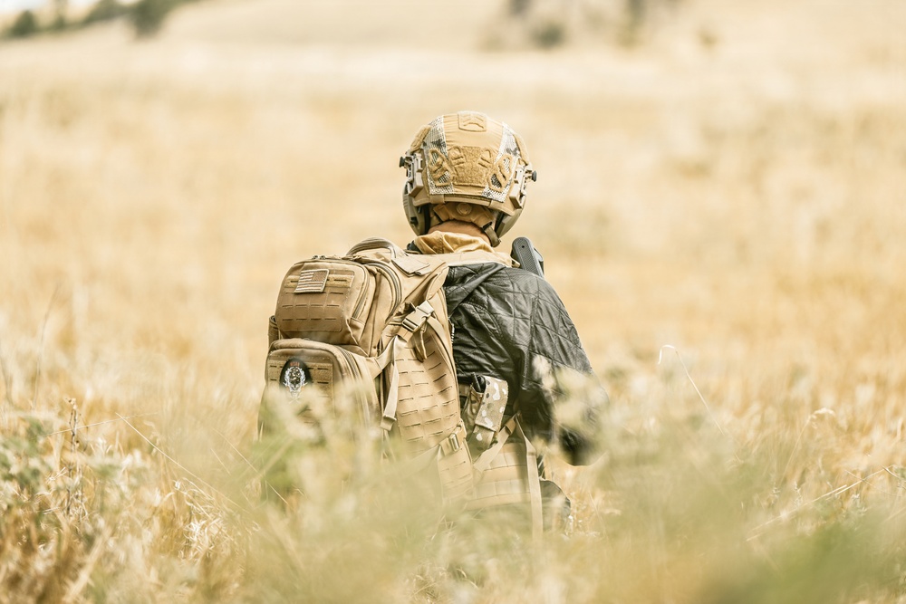 A person wearing full tactical military gear is seen from behind, moving through a dry grassland field. They are equipped with a tan/coyote brown tactical helmet, plate carrier vest, and a MOLLE-compatible backpack with a visible American flag patch. The warm, golden tones of the dry grass suggest an arid or semi-arid environment during late summer or autumn.