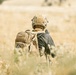 A person wearing full tactical military gear is seen from behind, moving through a dry grassland field. They are equipped with a tan/coyote brown tactical helmet, plate carrier vest, and a MOLLE-compatible backpack with a visible American flag patch. The warm, golden tones of the dry grass suggest an arid or semi-arid environment during late summer or autumn.
