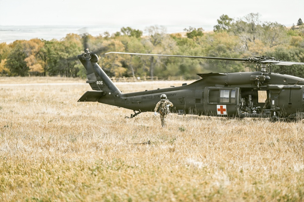 A United States Army medical evacuation (MEDEVAC) helicopter, tail number 605, is landed in a dry grassy field. A soldier in full combat gear is walking away from the aircraft toward the camera. The helicopter displays a prominent Red Cross symbol, identifying it as a medical support aircraft.