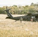 A United States Army medical evacuation (MEDEVAC) helicopter, tail number 605, is landed in a dry grassy field. A soldier in full combat gear is walking away from the aircraft toward the camera. The helicopter displays a prominent Red Cross symbol, identifying it as a medical support aircraft.