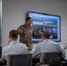 A U.S. Army soldier, identified by name tape as 'Alexander,' is presenting to a small group of civilians in a modern conference room. The presentation displayed on a large screen behind him is titled 'Civil Support Team' and appears to contain mission statistics and supporting imagery. The setting suggests a military briefing or outreach event to civilian stakeholders.
