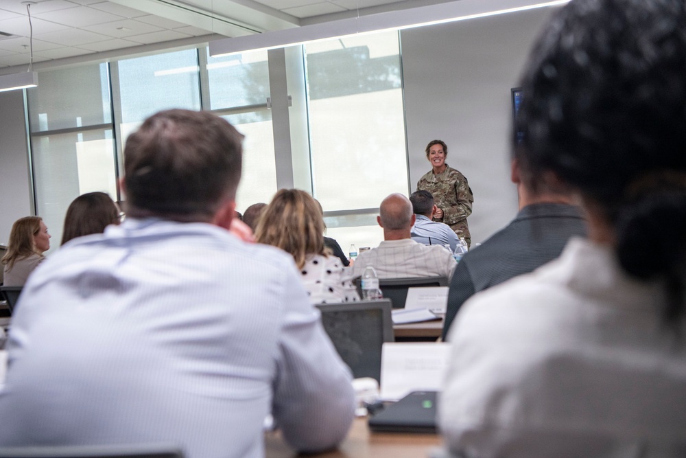 A person in military camouflage uniform is presenting or speaking at the front of a modern classroom or conference room. An audience of approximately 8-10 civilians is seated at tables facing the presenter, with documents, laptops, and water bottles visible on the tables. The setting appears to be a professional briefing or educational session.