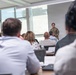A person in military camouflage uniform is presenting or speaking at the front of a modern classroom or conference room. An audience of approximately 8-10 civilians is seated at tables facing the presenter, with documents, laptops, and water bottles visible on the tables. The setting appears to be a professional briefing or educational session.