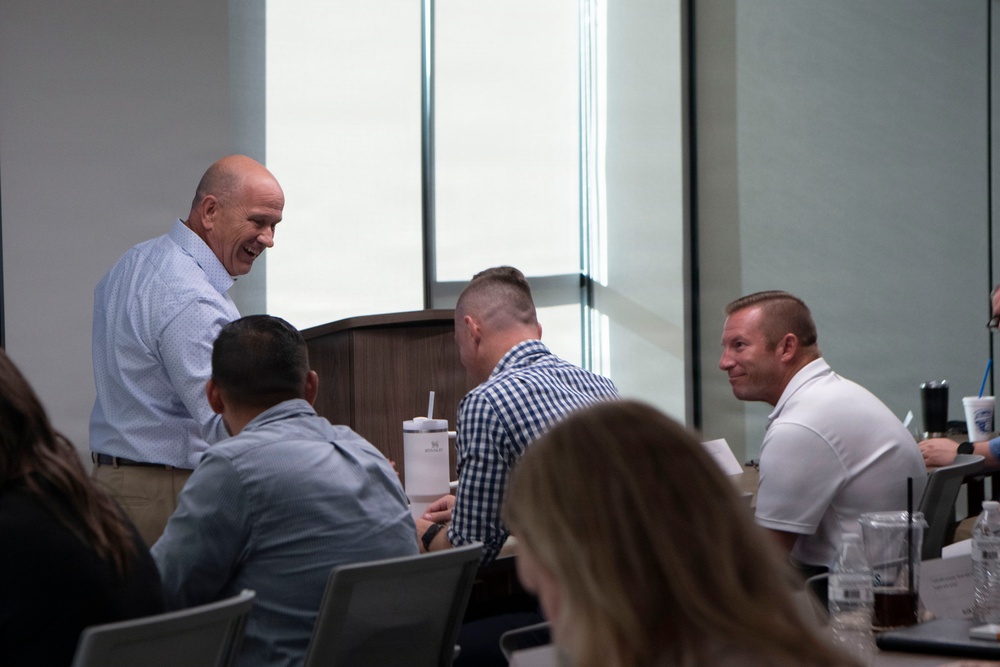 A group of people are gathered around a table in what appears to be a conference or meeting room setting. A tall, bald man in a light blue button-down shirt is standing and smiling, engaging with seated attendees. The atmosphere appears informal and conversational, suggesting a break or networking moment during a professional event.