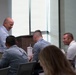 A group of people are gathered around a table in what appears to be a conference or meeting room setting. A tall, bald man in a light blue button-down shirt is standing and smiling, engaging with seated attendees. The atmosphere appears informal and conversational, suggesting a break or networking moment during a professional event.