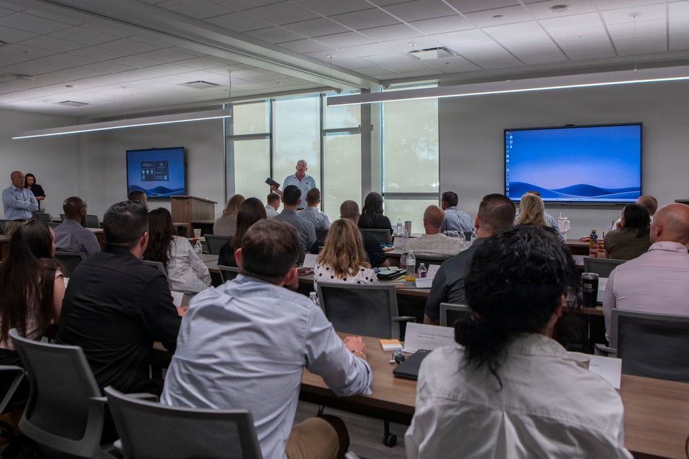 A professional training or corporate meeting is taking place in a modern conference room. A presenter stands at the front of the room addressing approximately 25-30 seated attendees arranged in rows at tables. Two large display screens are visible on either side of the room showing a Windows desktop interface.
