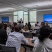 A professional training or corporate meeting is taking place in a modern conference room. A presenter stands at the front of the room addressing approximately 25-30 seated attendees arranged in rows at tables. Two large display screens are visible on either side of the room showing a Windows desktop interface.