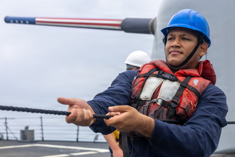 A sailor aboard what appears to be a U.S. Navy warship is handling a line (rope) during what appears to be an underway replenishment or mooring operation. The sailor is wearing standard naval working gear including a blue hard hat and red inflatable life vest. In the background, a naval gun barrel decorated with an American flag pattern is visible, along with another crew member in a white hard hat.