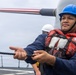 A sailor aboard what appears to be a U.S. Navy warship is handling a line (rope) during what appears to be an underway replenishment or mooring operation. The sailor is wearing standard naval working gear including a blue hard hat and red inflatable life vest. In the background, a naval gun barrel decorated with an American flag pattern is visible, along with another crew member in a white hard hat.
