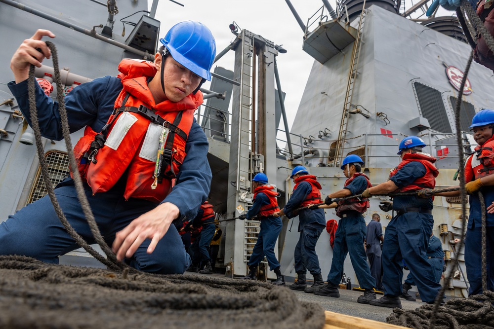 A group of naval personnel in blue coveralls, orange life vests, and blue hard hats are working on the deck of a warship handling thick mooring lines. The foreground sailor is coiling or managing a large rope while a line of crew members behind him appear to be hauling or tending a line in a coordinated effort. The vessel appears to be a U.S. Navy guided-missile destroyer based on the visible superstructure and hull design.