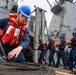 A group of naval personnel in blue coveralls, orange life vests, and blue hard hats are working on the deck of a warship handling thick mooring lines. The foreground sailor is coiling or managing a large rope while a line of crew members behind him appear to be hauling or tending a line in a coordinated effort. The vessel appears to be a U.S. Navy guided-missile destroyer based on the visible superstructure and hull design.