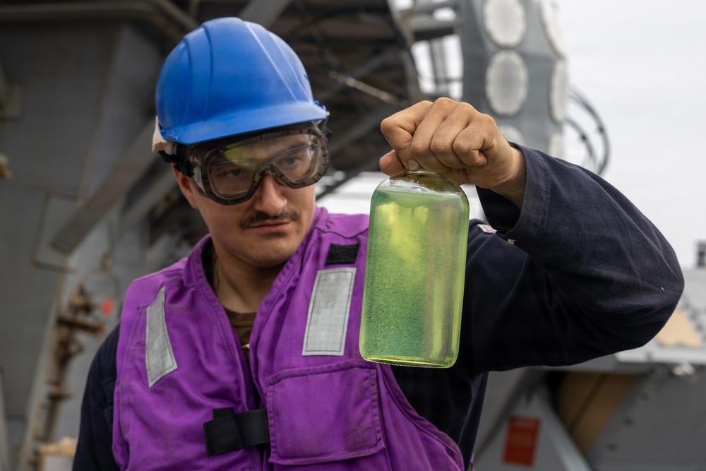 A worker wearing a blue hard hat, safety goggles, and a purple safety vest holds up a clear glass jar containing a yellow-green liquid for inspection. The setting appears to be the deck of a naval or industrial vessel, with metal structures and equipment visible in the background. The individual appears to be conducting a fuel or fluid quality check.