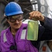 A worker wearing a blue hard hat, safety goggles, and a purple safety vest holds up a clear glass jar containing a yellow-green liquid for inspection. The setting appears to be the deck of a naval or industrial vessel, with metal structures and equipment visible in the background. The individual appears to be conducting a fuel or fluid quality check.