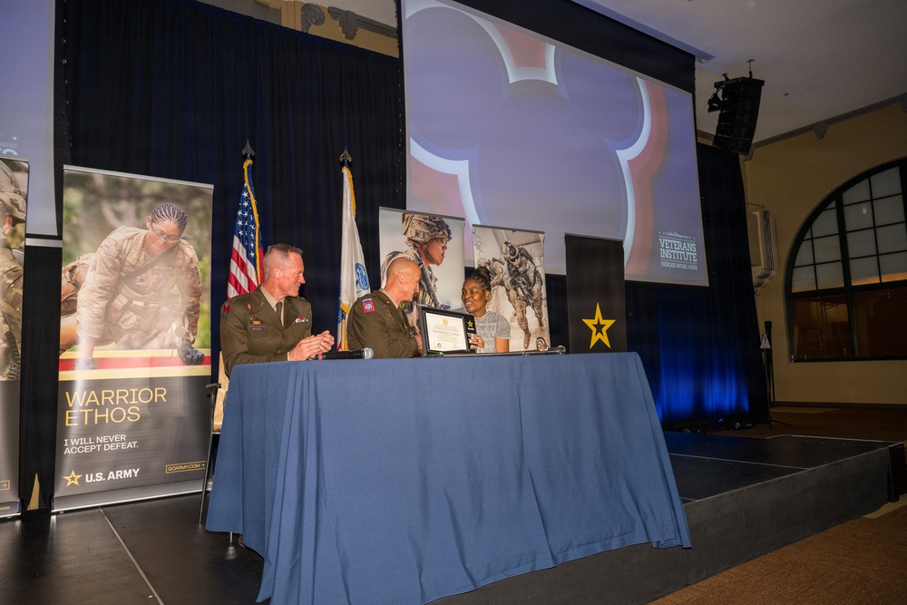 A formal ceremony is taking place on a stage, likely a military recognition or award event co-hosted by the U.S. Army and Disney's Veterans Institute. Two uniformed U.S. Army officers are presenting a framed certificate or award to a civilian woman at a blue-draped table. The backdrop includes a projected Mickey Mouse silhouette and a 'Veterans Institute - Heroes Work Here' logo.