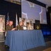 A formal ceremony is taking place on a stage, likely a military recognition or award event co-hosted by the U.S. Army and Disney's Veterans Institute. Two uniformed U.S. Army officers are presenting a framed certificate or award to a civilian woman at a blue-draped table. The backdrop includes a projected Mickey Mouse silhouette and a 'Veterans Institute - Heroes Work Here' logo.