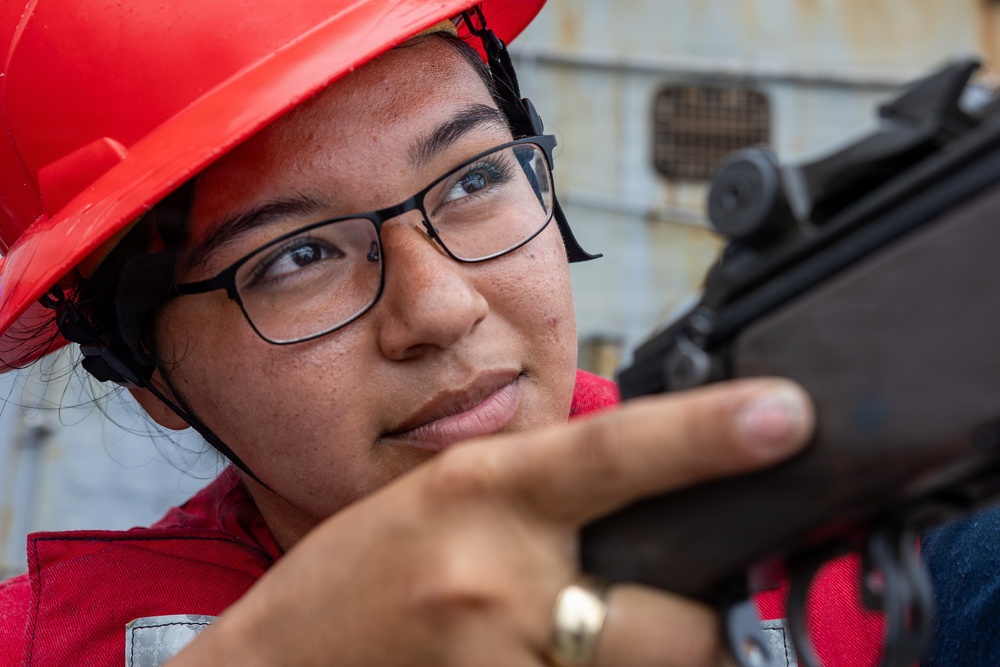 A young woman wearing a red hard hat and black-framed glasses is closely examining or aiming a firearm, likely a rifle or carbine. She appears to be in a training or inspection context, wearing red safety/work attire. The close-up shot captures her focused expression as she looks down the sights of the weapon.