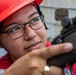 A young woman wearing a red hard hat and black-framed glasses is closely examining or aiming a firearm, likely a rifle or carbine. She appears to be in a training or inspection context, wearing red safety/work attire. The close-up shot captures her focused expression as she looks down the sights of the weapon.