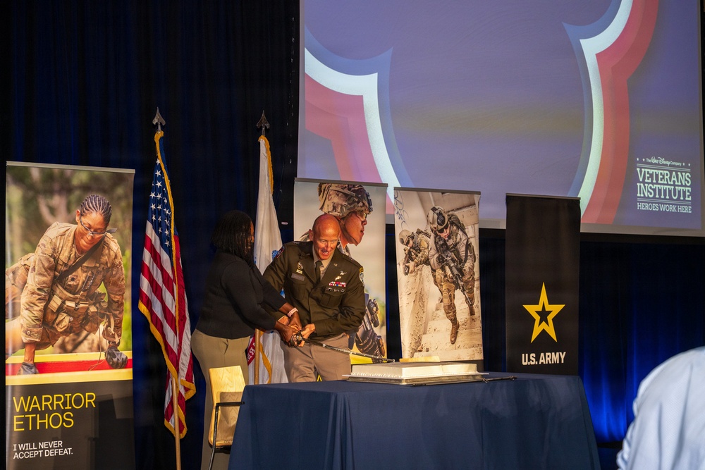 A military ceremony or event appears to be taking place at a joint U.S. Army and Walt Disney Company Veterans Institute event. A uniformed Army soldier and a civilian woman are cutting what appears to be a ceremonial cake on a blue-draped table. The setting includes patriotic banners, U.S. and military flags, and recruitment/promotional imagery.