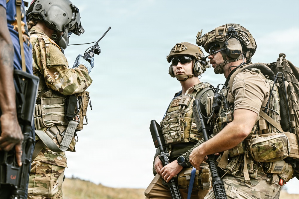 Three heavily equipped military or law enforcement personnel are gathered in an outdoor field environment. One individual is operating a handheld radio while the others appear to be listening or awaiting instructions. All personnel are wearing tactical gear, helmets, and body armor consistent with special operations or EOD (Explosive Ordnance Disposal) units.