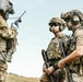 Three heavily equipped military or law enforcement personnel are gathered in an outdoor field environment. One individual is operating a handheld radio while the others appear to be listening or awaiting instructions. All personnel are wearing tactical gear, helmets, and body armor consistent with special operations or EOD (Explosive Ordnance Disposal) units.