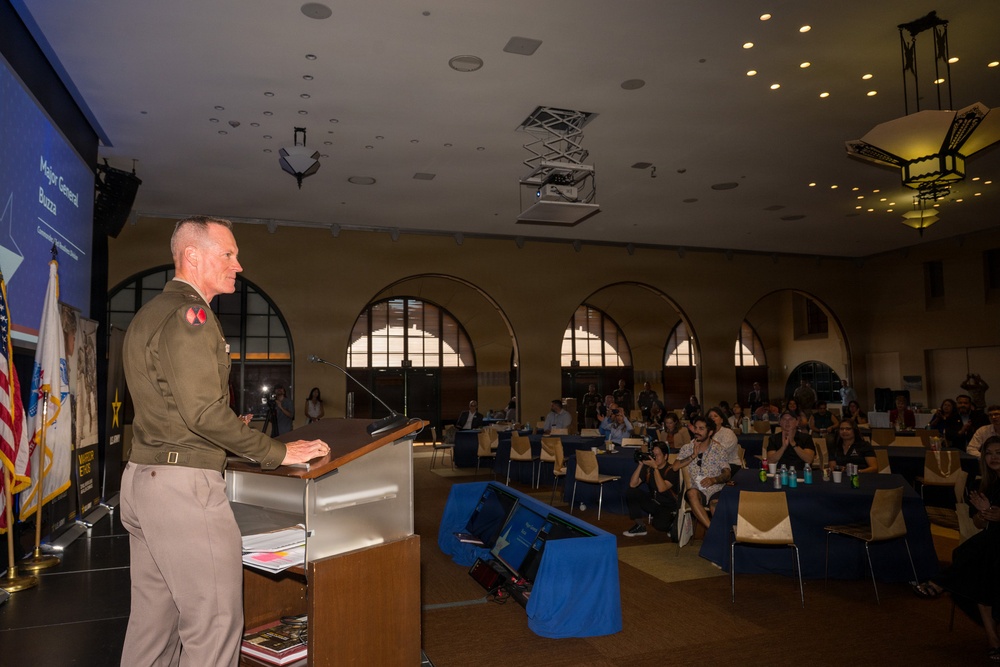 A U.S. Army Major General identified on the presentation screen as 'Major General Buzz' is delivering a speech at a podium during a formal conference or symposium. The officer is in military service uniform addressing a seated audience in a large, architecturally distinguished hall with arched windows. The event appears to be a professional or public engagement event with civilian attendees.