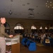 A U.S. Army Major General identified on the presentation screen as 'Major General Buzz' is delivering a speech at a podium during a formal conference or symposium. The officer is in military service uniform addressing a seated audience in a large, architecturally distinguished hall with arched windows. The event appears to be a professional or public engagement event with civilian attendees.