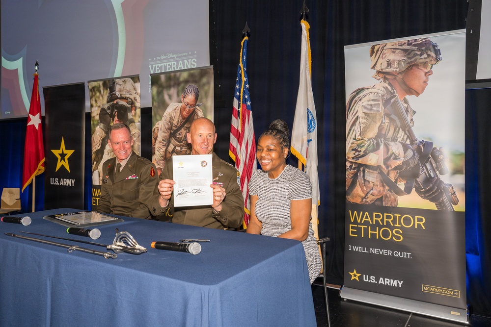 A formal U.S. Army ceremony appears to be taking place, likely at a Walt Disney Company Veterans event. Two individuals in U.S. Army uniforms and one civilian woman are seated at a table, holding up a signed official document. The setting includes U.S. Army branding, recruitment banners, American flags, and military regalia.
