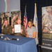 A formal U.S. Army ceremony appears to be taking place, likely at a Walt Disney Company Veterans event. Two individuals in U.S. Army uniforms and one civilian woman are seated at a table, holding up a signed official document. The setting includes U.S. Army branding, recruitment banners, American flags, and military regalia.