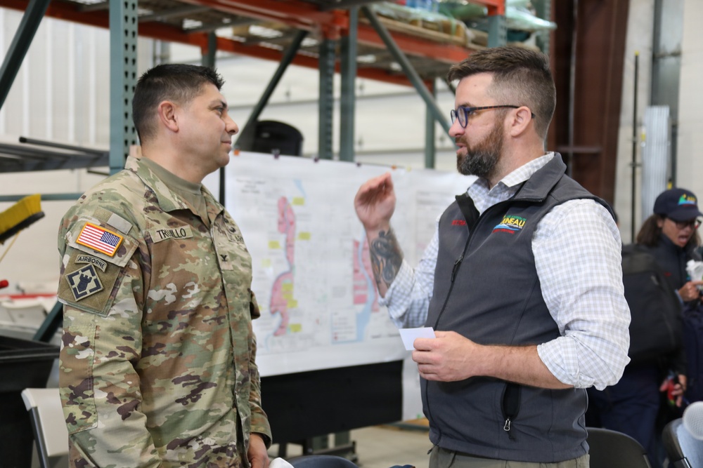 A U.S. Army soldier in camouflage uniform is engaged in a conversation with a civilian male inside what appears to be a warehouse or operations center. A large map or planning document is visible on the wall behind them, suggesting a coordination or briefing context. The setting appears to be an emergency management or military planning environment.