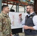 A U.S. Army soldier in camouflage uniform is engaged in a conversation with a civilian male inside what appears to be a warehouse or operations center. A large map or planning document is visible on the wall behind them, suggesting a coordination or briefing context. The setting appears to be an emergency management or military planning environment.