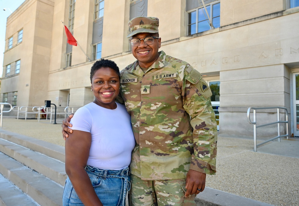 A U.S. Army Sergeant named Blocker in OCP (Operational Camouflage Pattern) uniform poses with a civilian woman in front of what appears to be a National Guard Armory building. Both individuals are smiling broadly, suggesting a celebratory or reunion moment. The soldier has his arm around the woman in a warm, familiar pose.